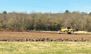 Yellow bulldozer on dirt field near trees under blue sky.
