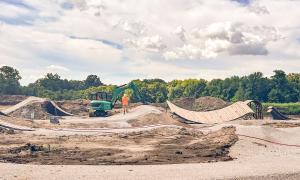 Construction site with a digger, dirt mounds, and trees in the background.